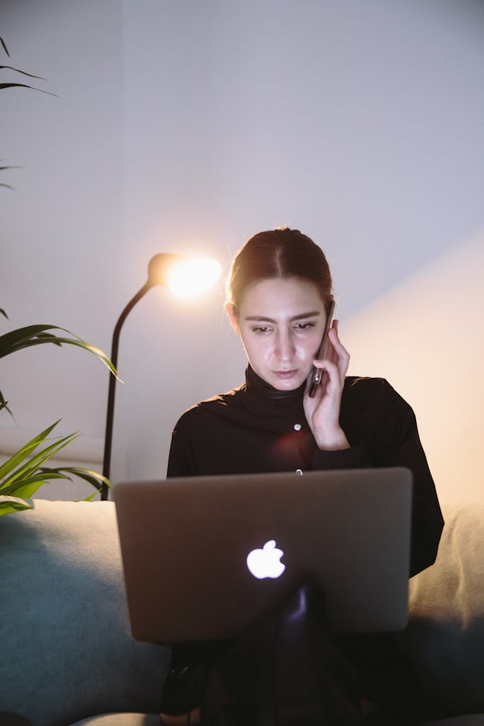 Focused woman using laptop and smartphone in a softly lit room for remote work.